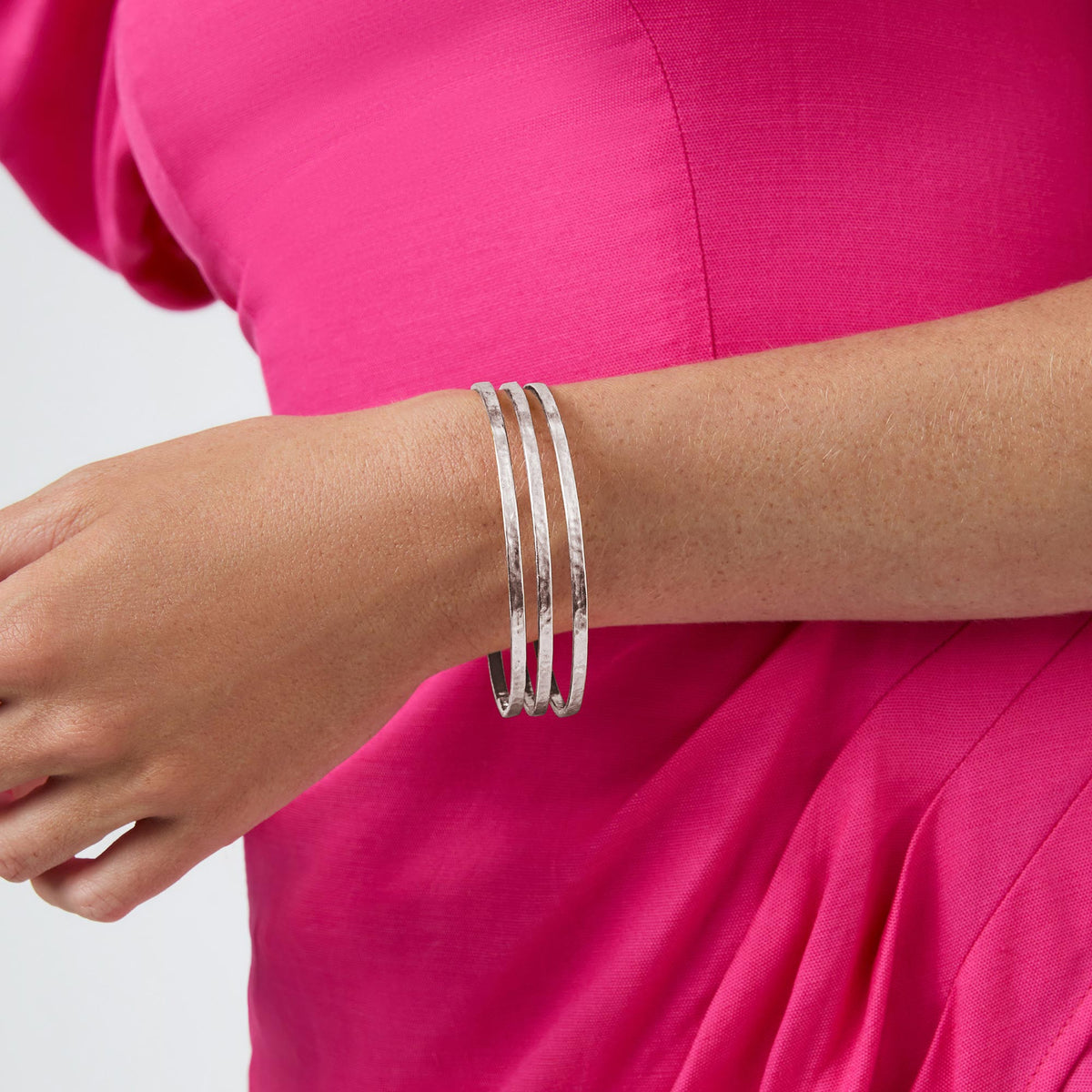 A person wearing three thin, textured silver Crescent Bangles on their wrist against a pink dress.