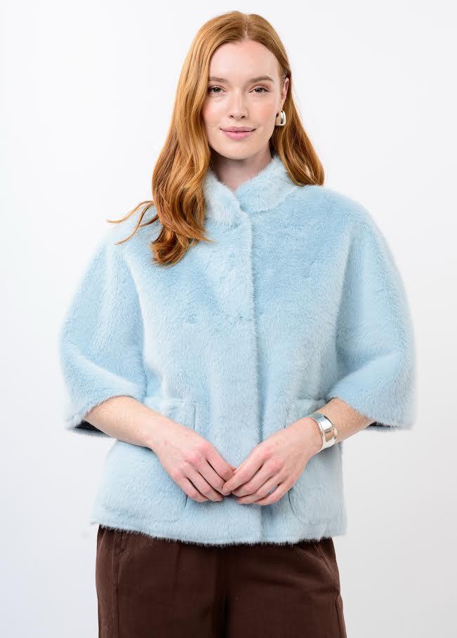 A woman wearing a light blue Megan Faux Fur Capelet with short sleeves and brown trousers.