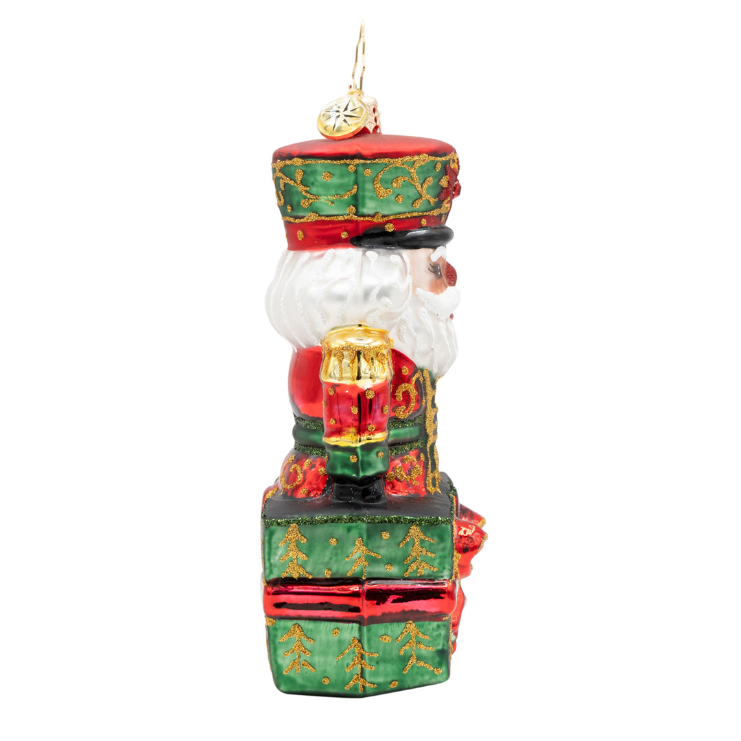 Glass nutcracker ornament in a red and green uniform sitting on a green gift box with gold trees.