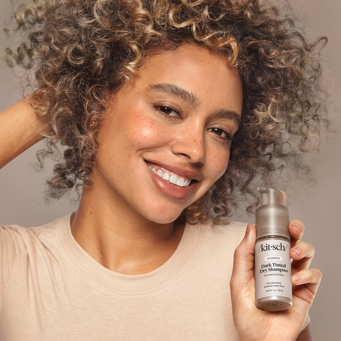 A woman with curly hair holds a bottle of Kitsch Dark Tinted Volumizing Dry Shampoo.