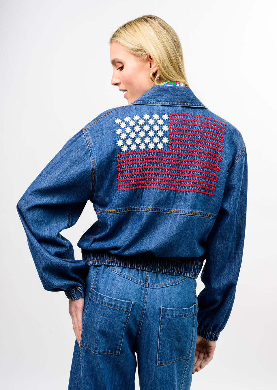 Back view of a blue denim bomber jacket with an embroidered American flag made of white flowers.