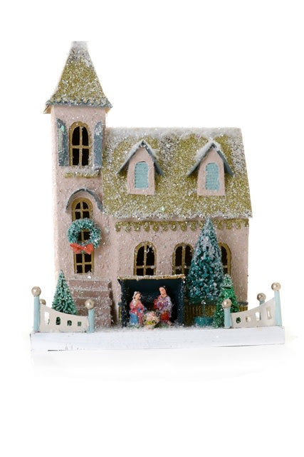 A miniature pink and green glittered church with a nativity scene inside, surrounded by snow and a white picket fence.