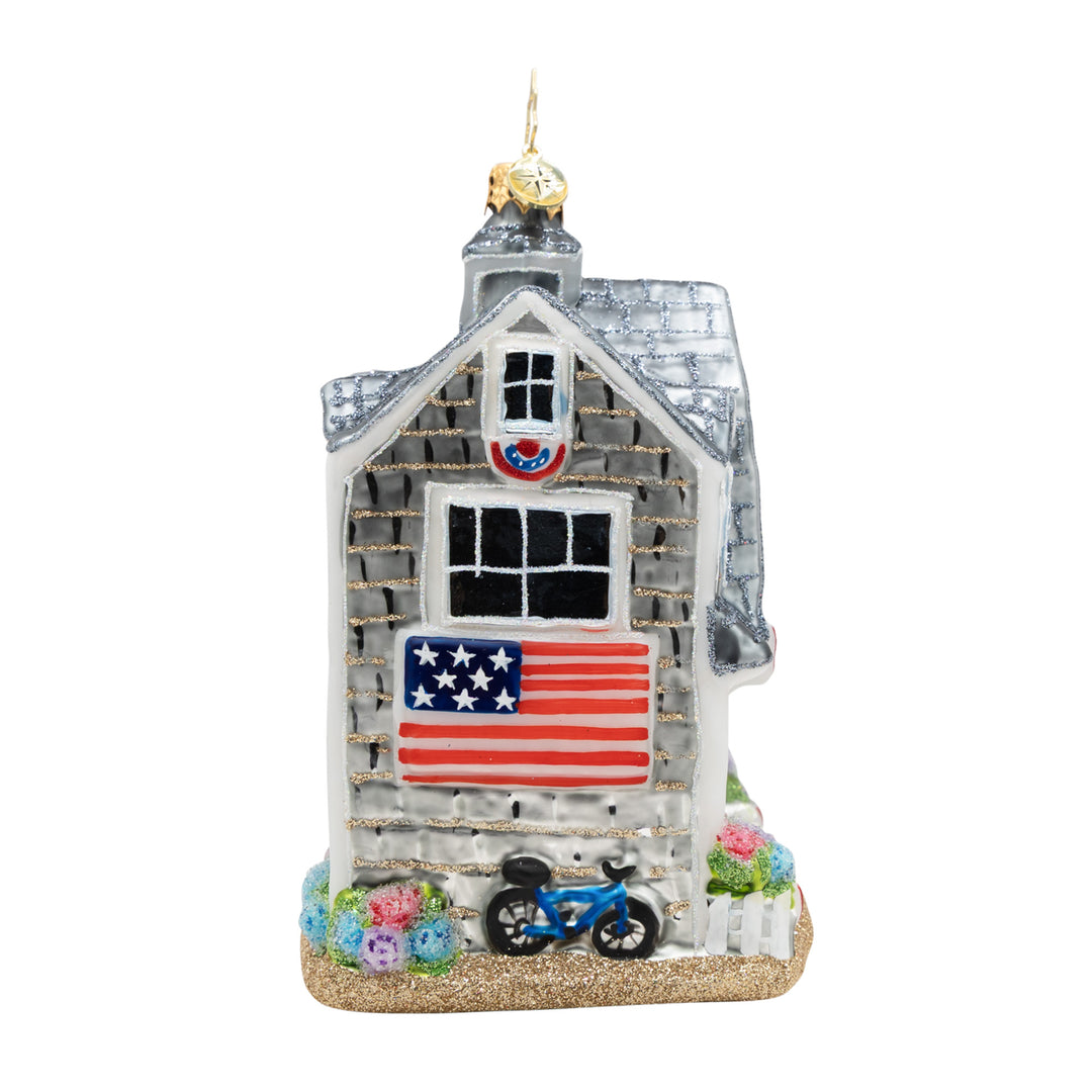 Glass beach house ornament with an American flag, bicycle, and colorful flowers on a glitter base.