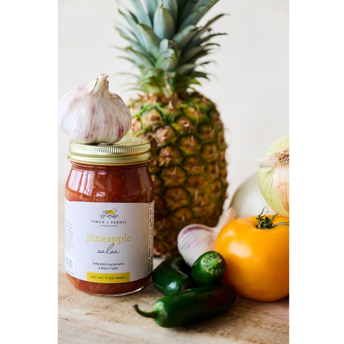 17 oz. jar of Finch + Fennel Pineapple Salsa surrounded by fresh pineapple, garlic, and peppers.