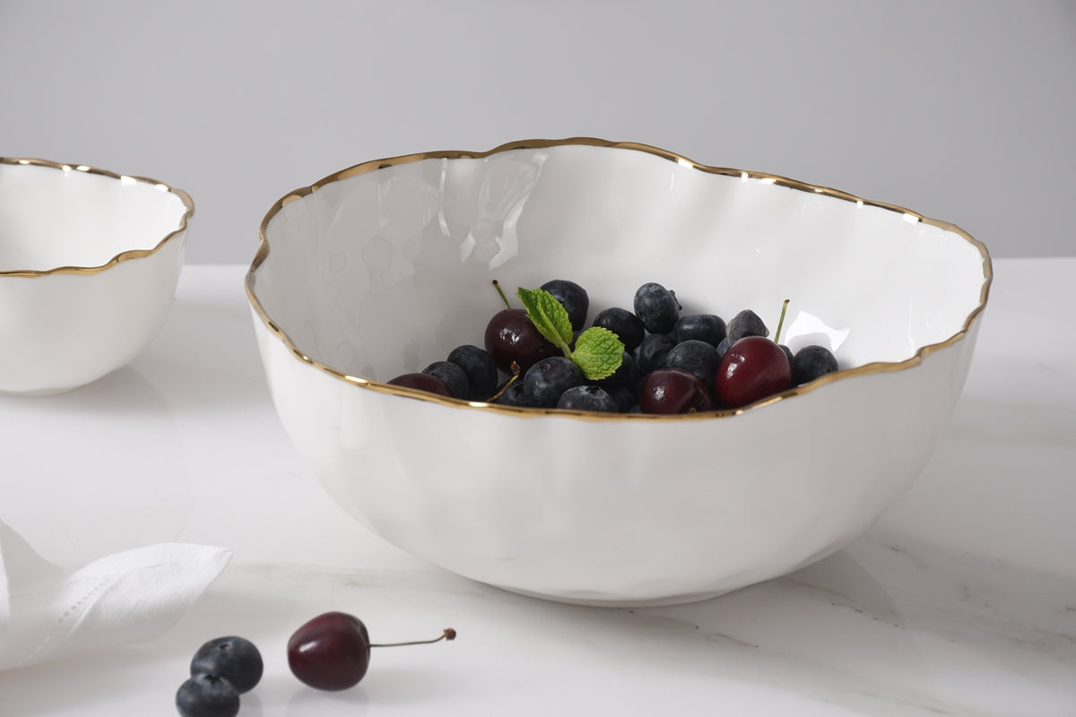 Medium white porcelain bowl with a gold rim, filled with blueberries and cherries on a white table.