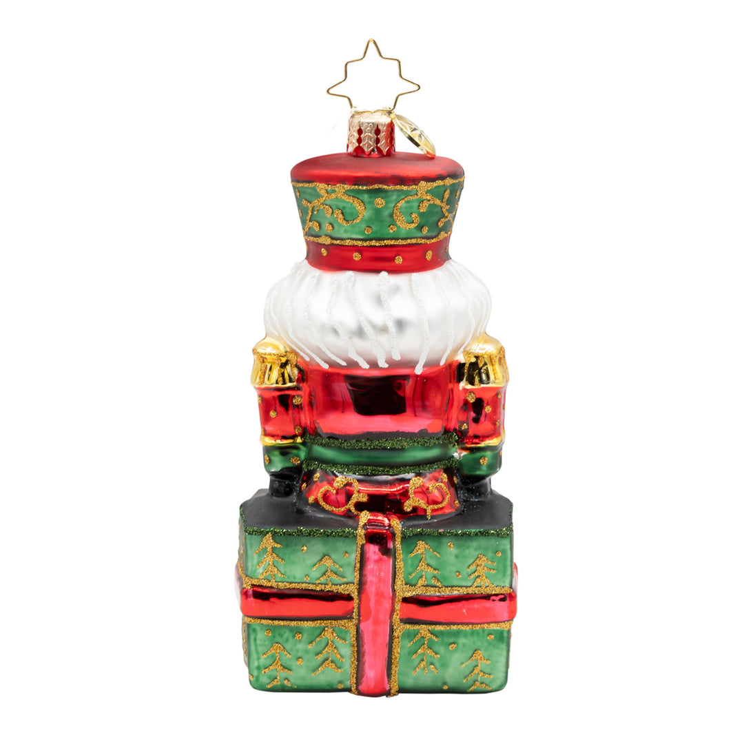 Glass nutcracker ornament in red and green sitting on a gift box with gold glitter accents.