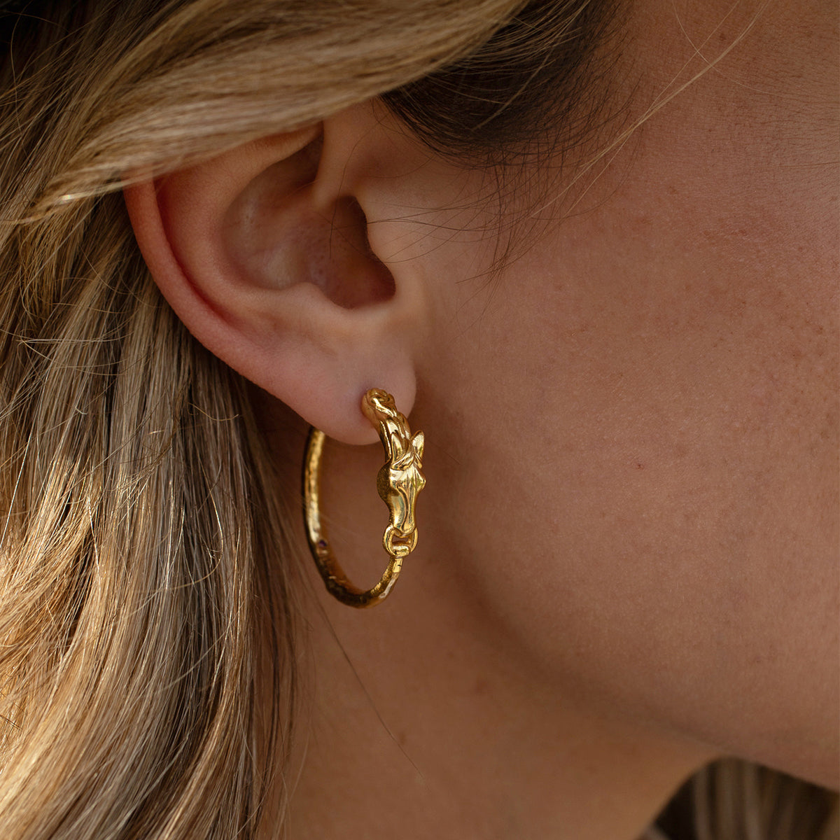 Close-up of a person wearing a gold equestrian hoop earring featuring a sculpted horsehead motif.