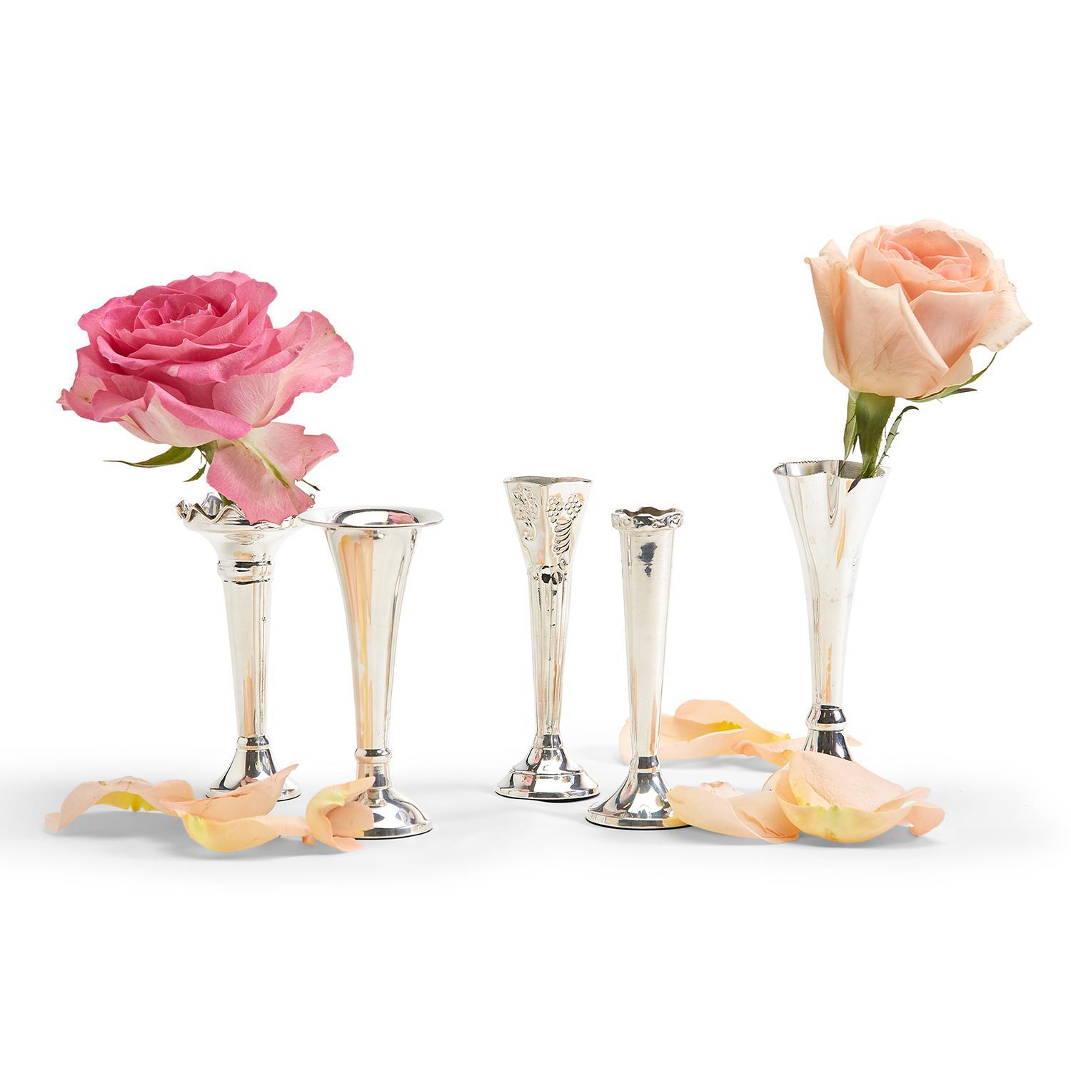 Five assorted silver-plated single stem vases of varying designs with two roses and loose petals.
