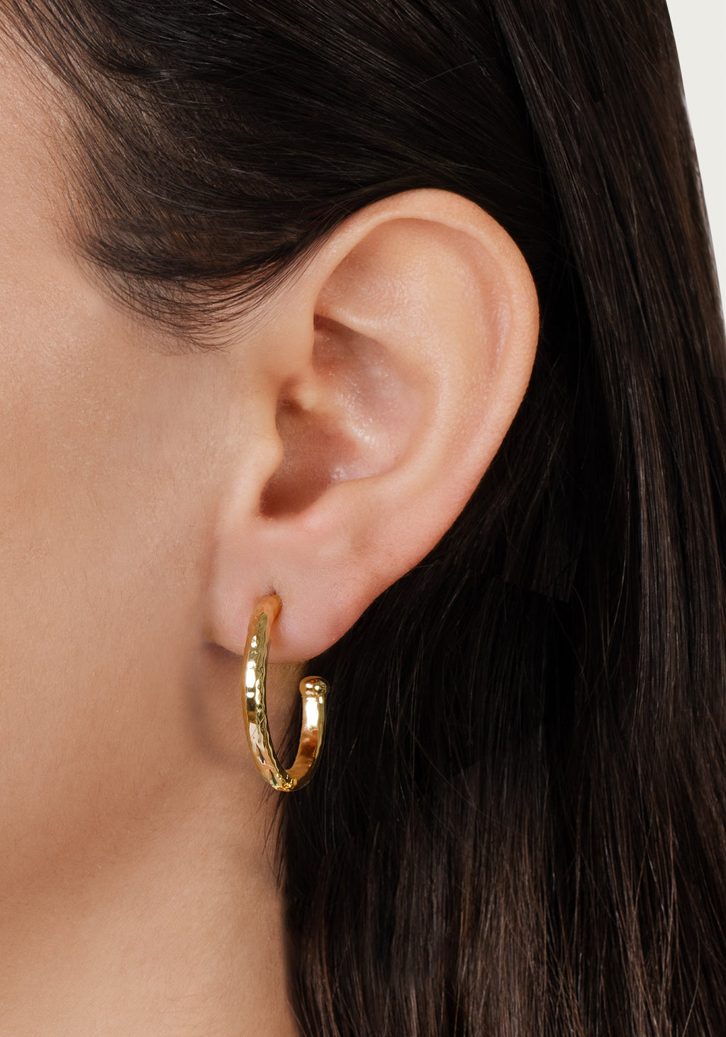 Close-up of a person wearing a medium-sized, textured gold Farrier Charm Hoop earring.