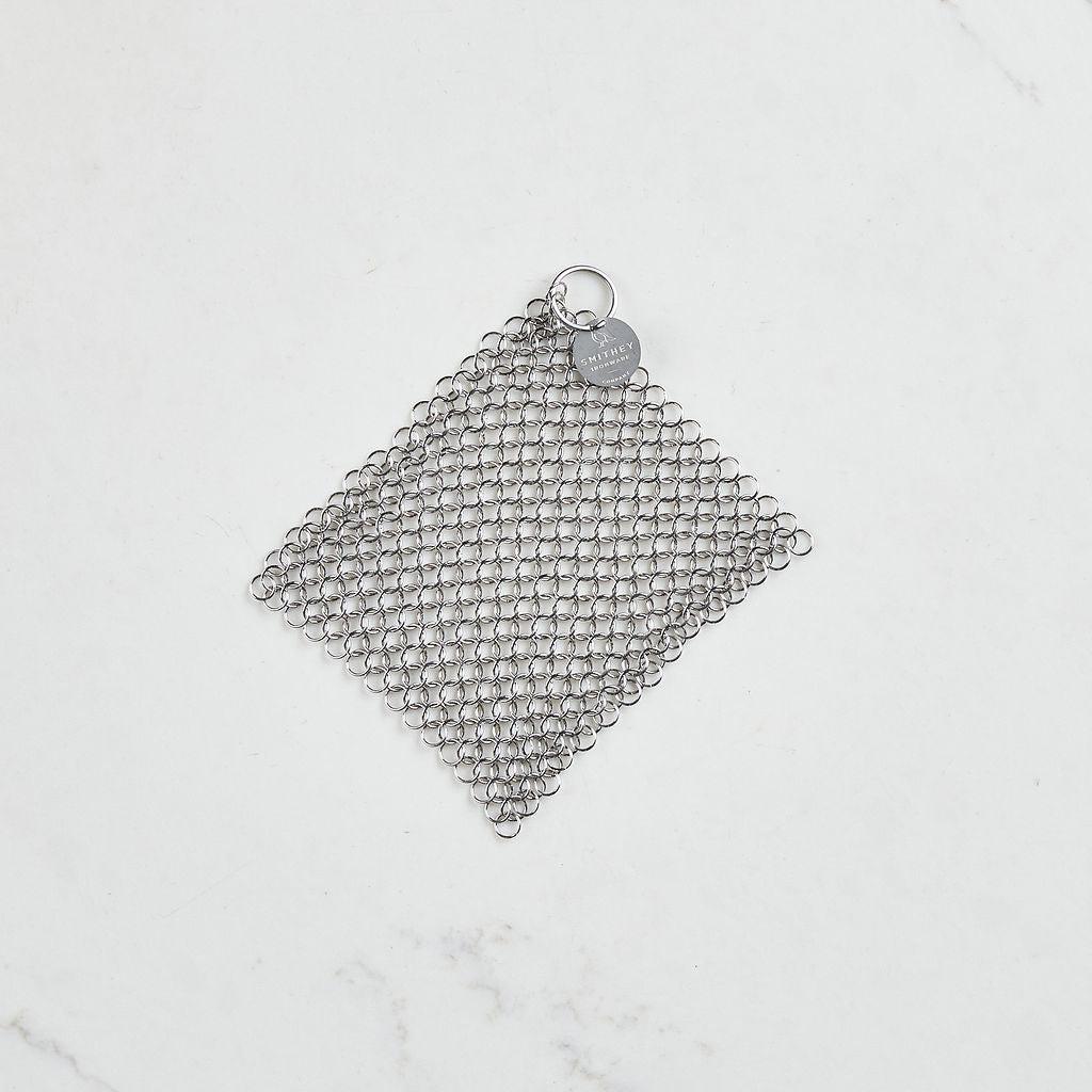 A 5x5 inch stainless steel chainmail scrubber with a circular logo tag on a white surface.