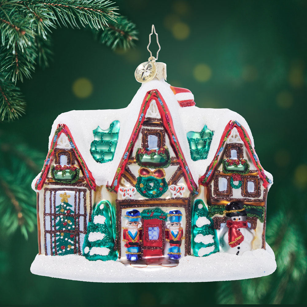 Glass Christmas ornament shaped like a snow-covered house with nutcrackers, a snowman, and a Christmas tree.