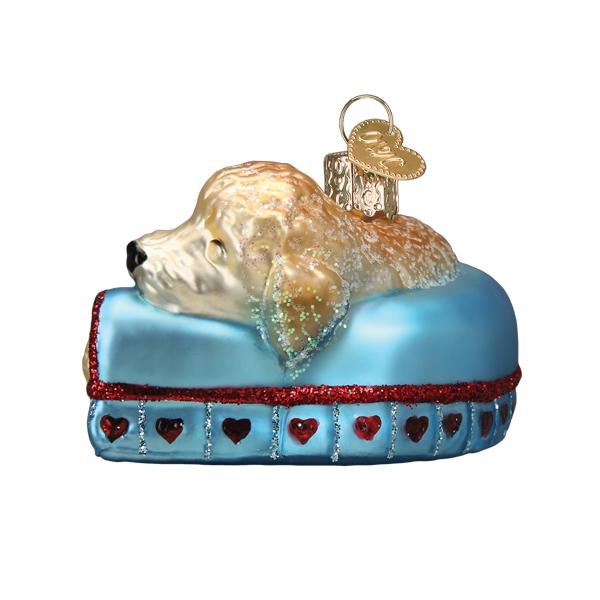 Glass Christmas ornament of a golden doodle dog sleeping in a blue bed with red glitter hearts.