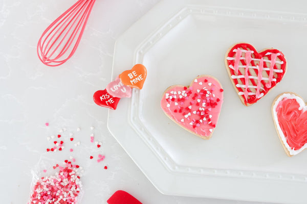 Heart-shaped sugar cookies with pink and red icing on a white plate next to a pink whisk.