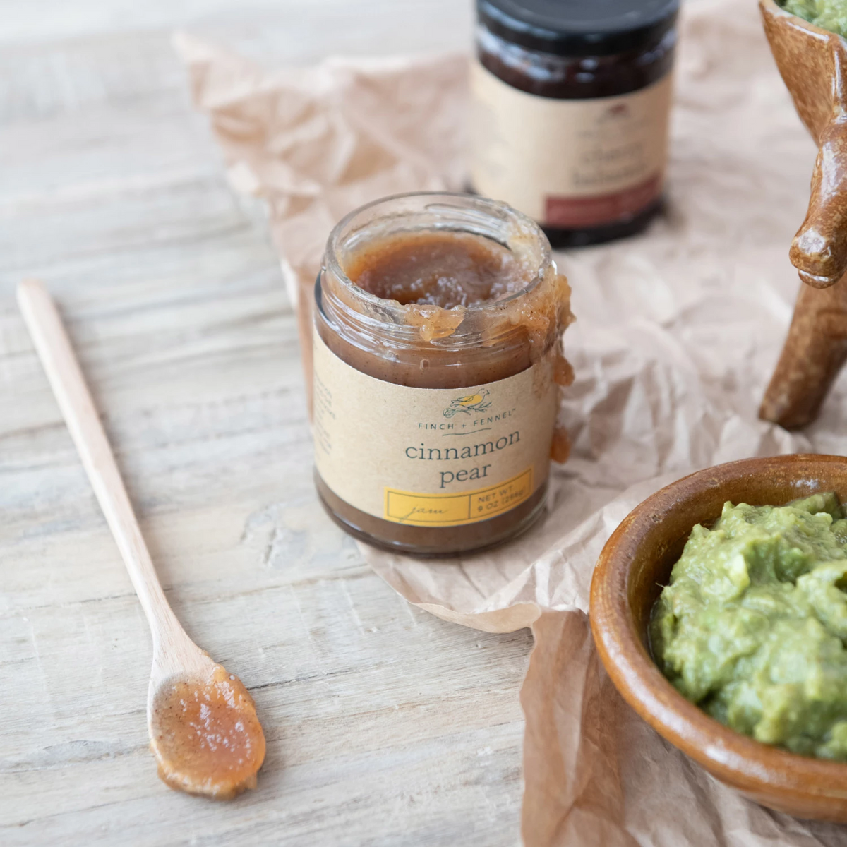 An open jar of Finch + Fennel Cinnamon Pear Jam next to a wooden spoon and a bowl of green spread.