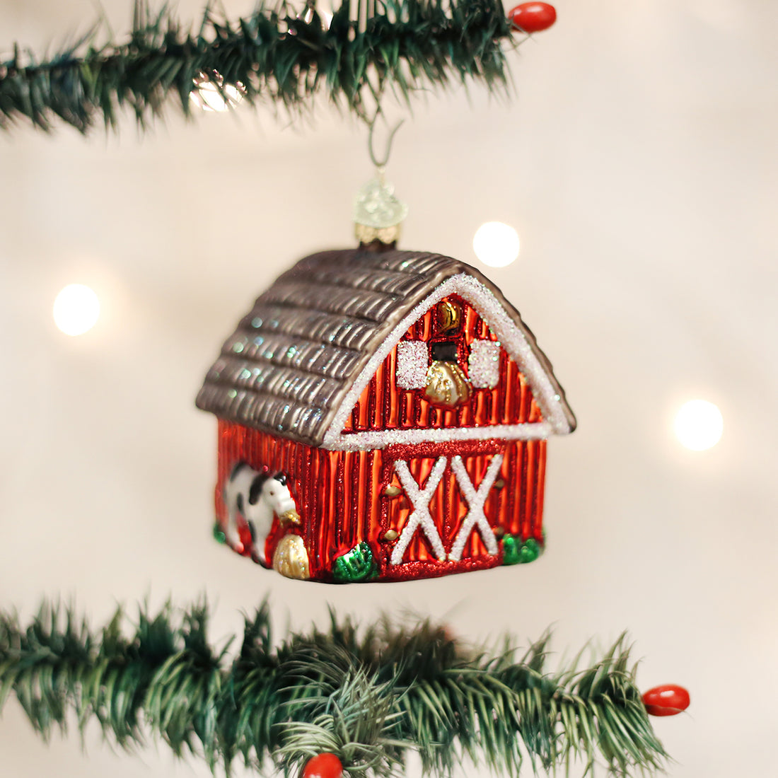 Red glass barn Christmas ornament with a brown roof and a cow detail hanging on a tree.