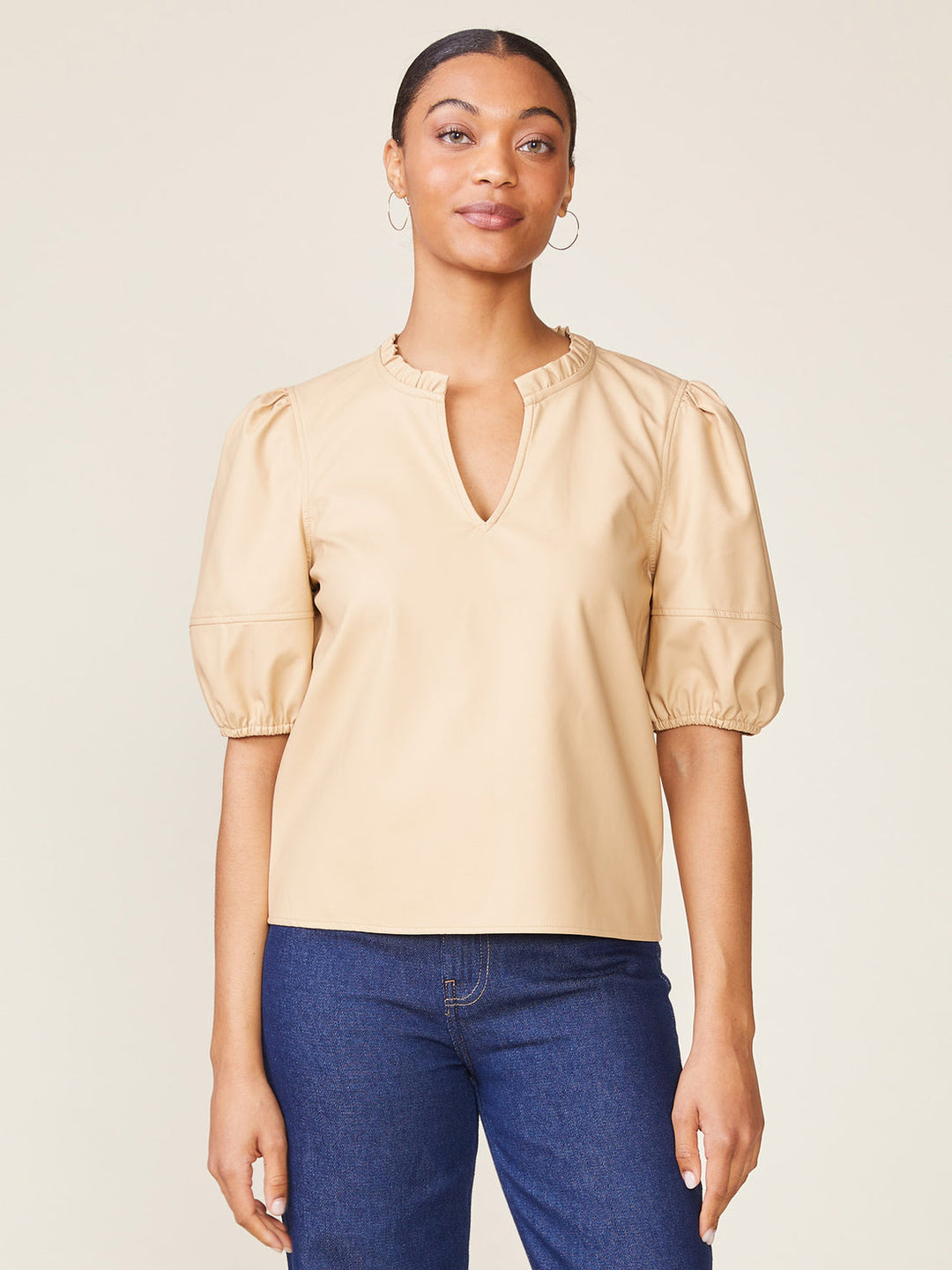 Woman wearing a beige Kacey Vegan Leather Blouse with puff sleeves and a ruffled split neckline.