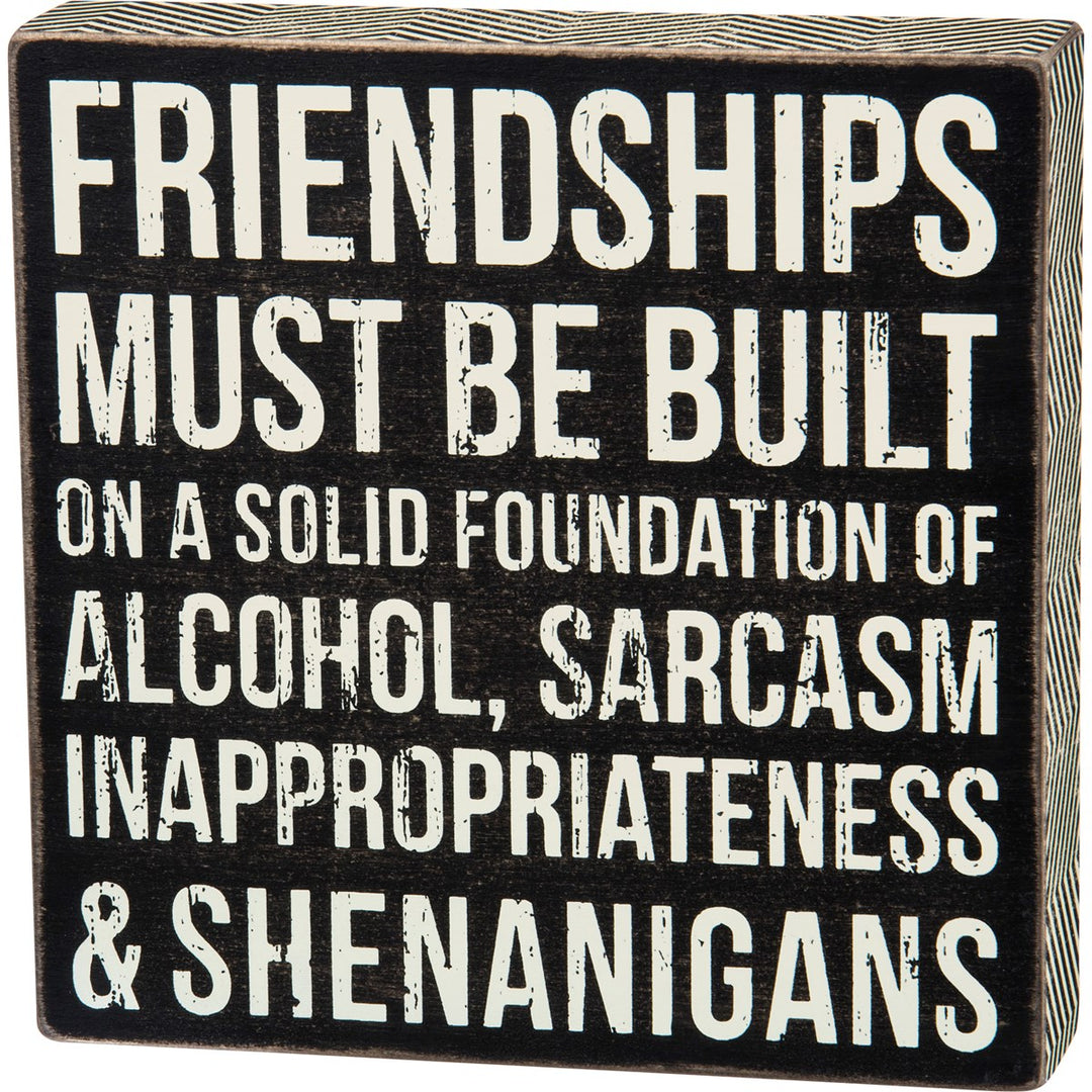 Black wooden box sign with white distressed text reading "Friendships must be built on a solid foundation of alcohol, sarcasm, inappropriateness & shenanigans."