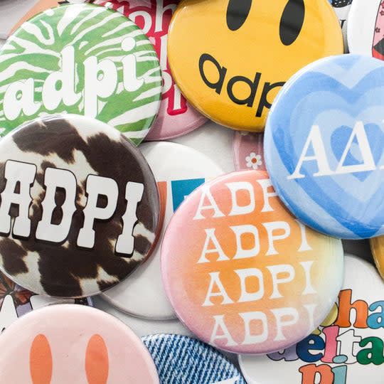 Assorted sorority pin-back buttons in various colors with Greek letter designs.