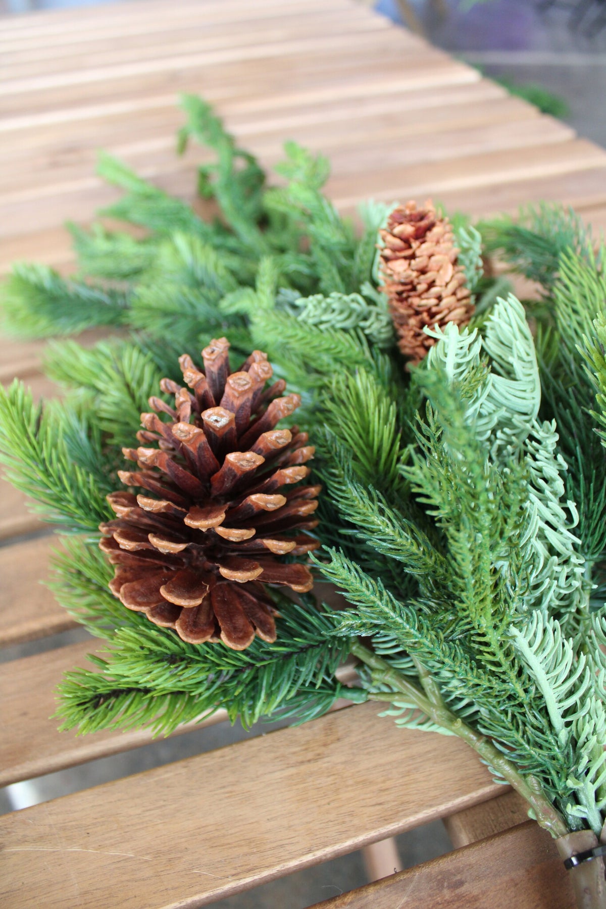 Artificial Norfolk spruce and juniper teardrop swag with two brown pinecones on a wooden surface.