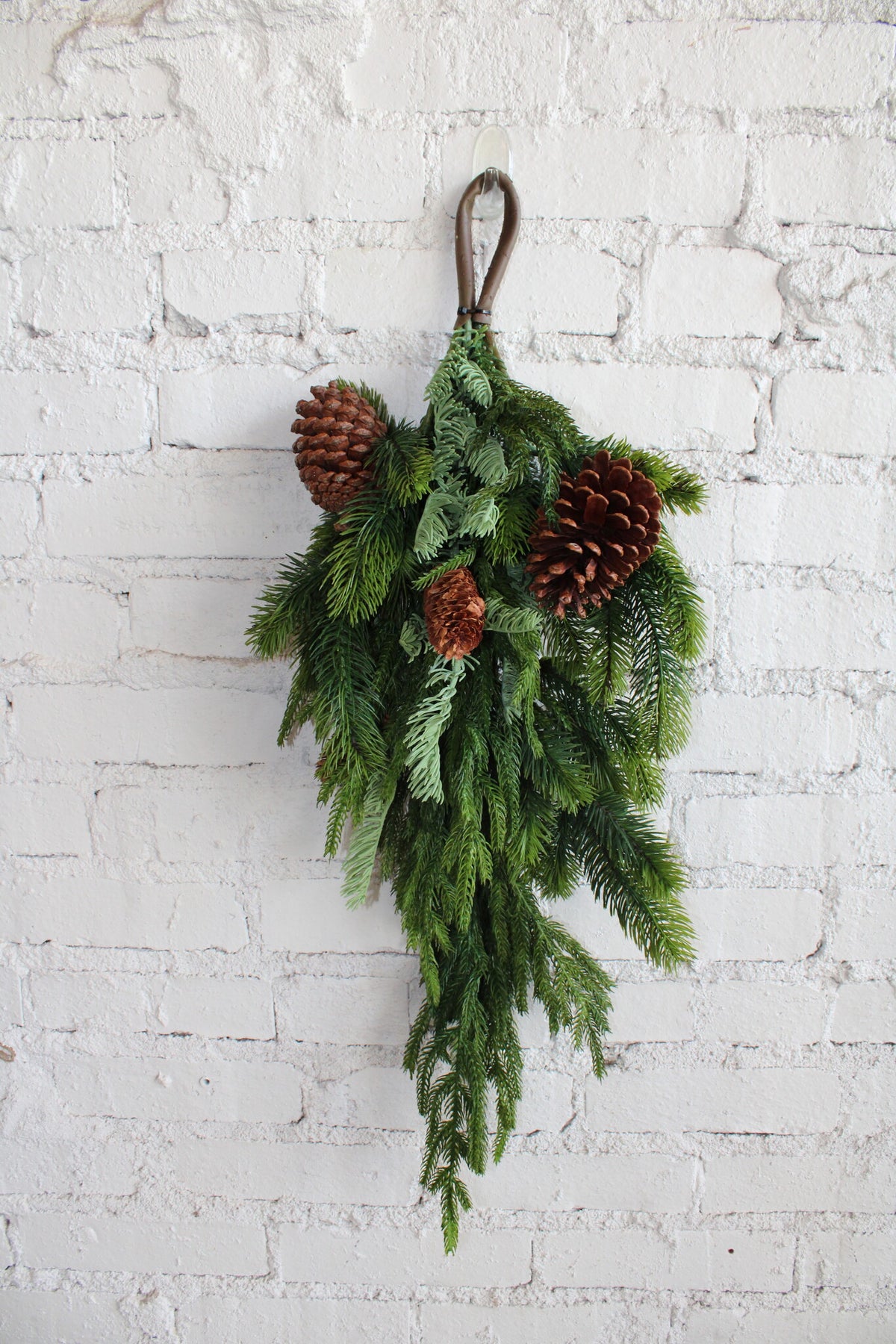 27-inch artificial Norfolk, spruce, and juniper teardrop swag with pinecones on a white brick wall.