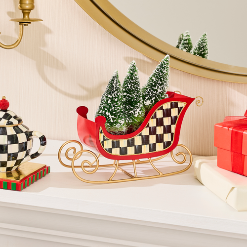 A red and checkered tin sleigh with gold runners holding three snow-dusted miniature pine trees.