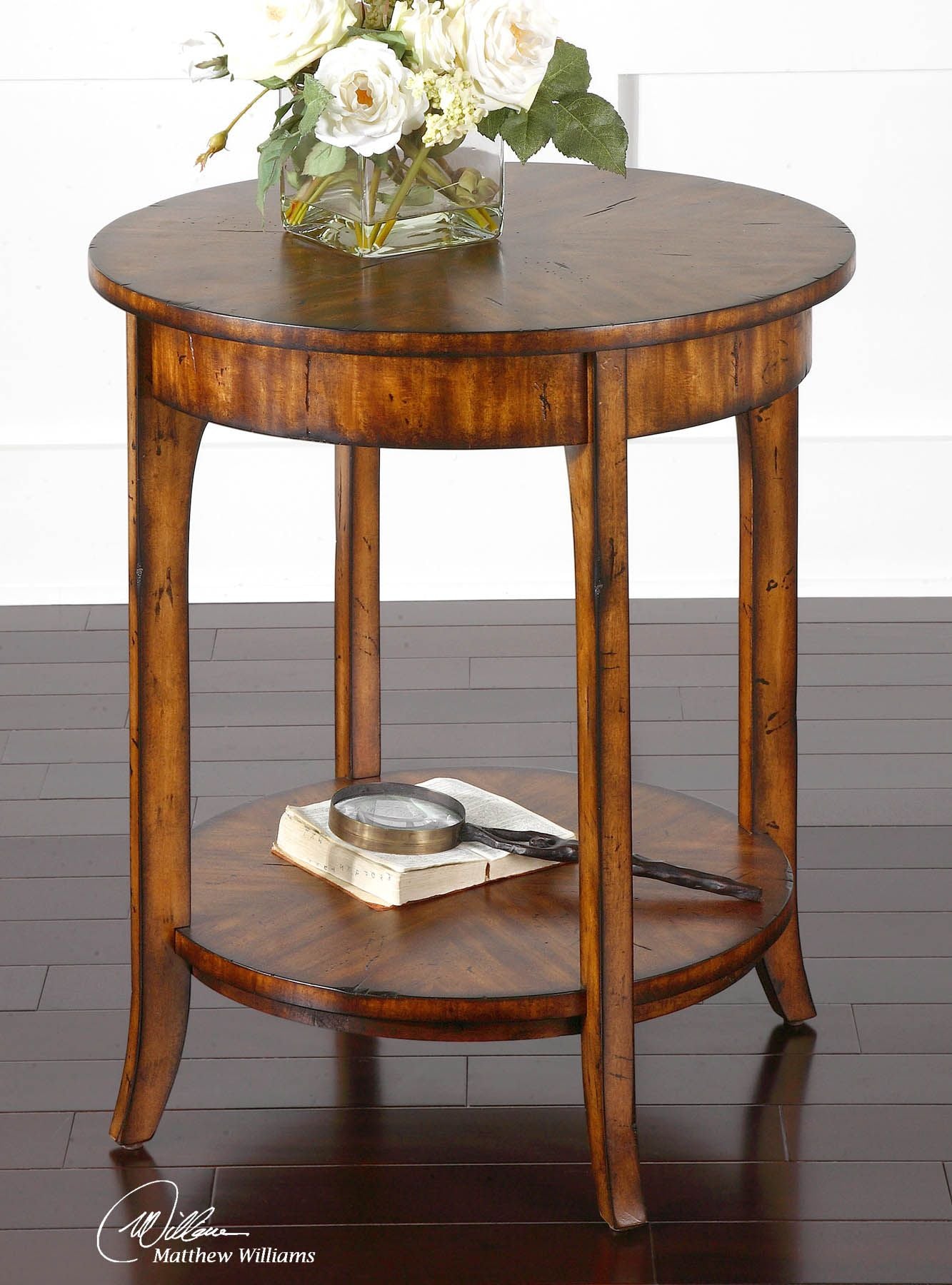 Round wooden end table with a lower shelf, featuring a dark wood finish and carved legs. A vase of white flowers sits on top.