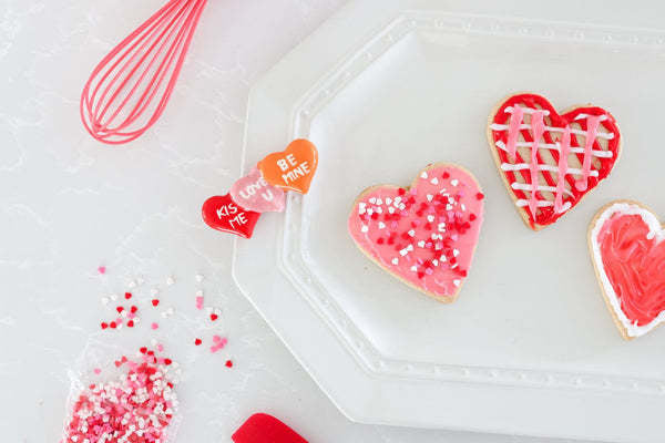 Heart-shaped sugar cookies with pink and red icing and sprinkles on a white scalloped plate.