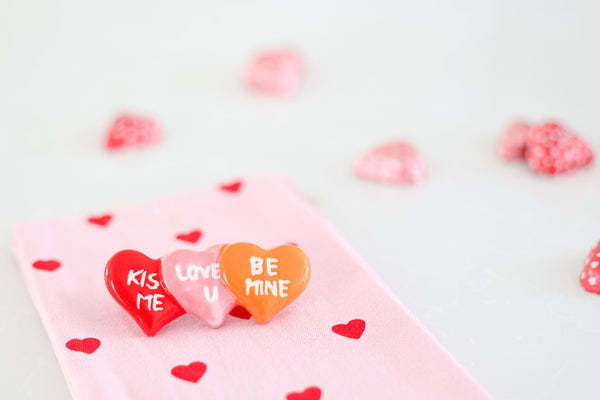 Three heart-shaped pins reading KISS ME, LOVE U, and BE MINE on a pink fabric with red heart print.