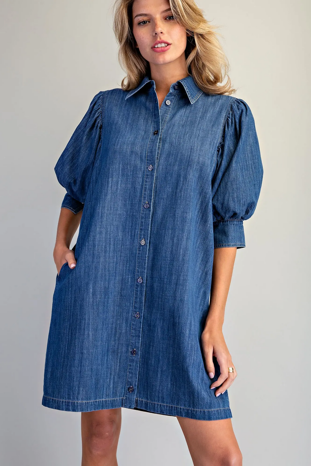 Woman wearing a blue denim button-down mini dress with puff sleeves and side pockets.