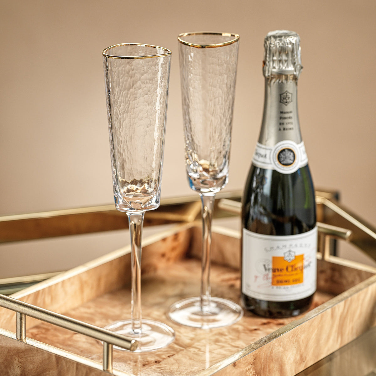 Two Aperitivo triangular champagne flutes with gold rims next to a bottle on a wooden tray.