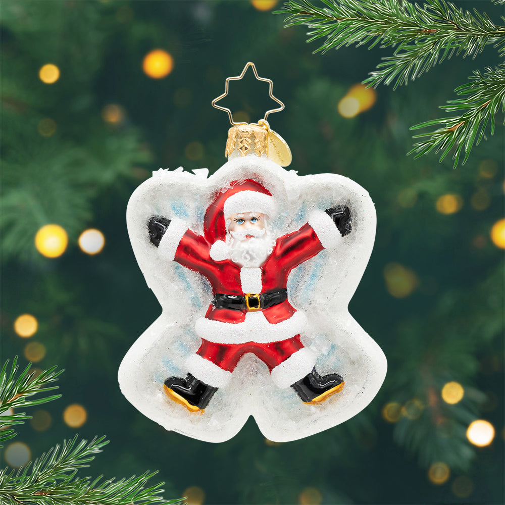 Glass Christmas ornament of Santa Claus making a snow angel, set against a blurred tree background.