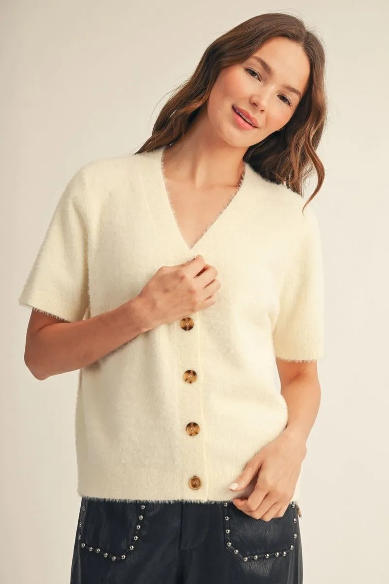 Woman wearing a cream-colored, short-sleeved fuzzy button-down cardigan with brown buttons.