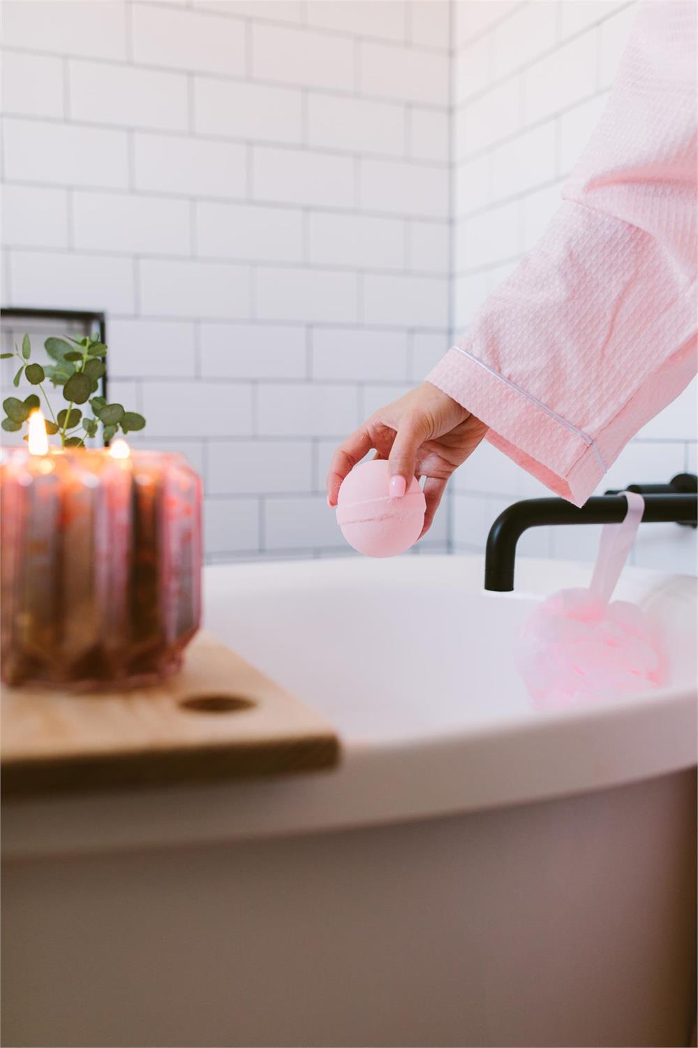 A hand holding a pink Sweet Grace Bath Bomb over a white bathtub with a pink robe and lit candles.