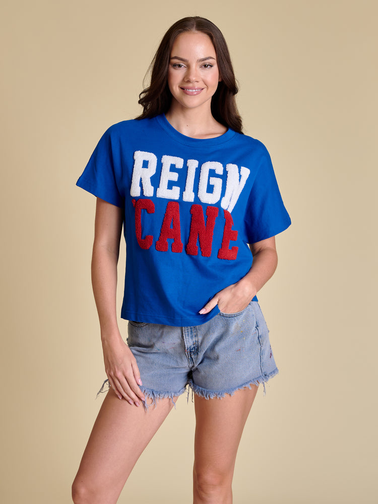 Woman wearing a blue boxy t-shirt with white and red chenille 'REIGN CANE' text and denim shorts.