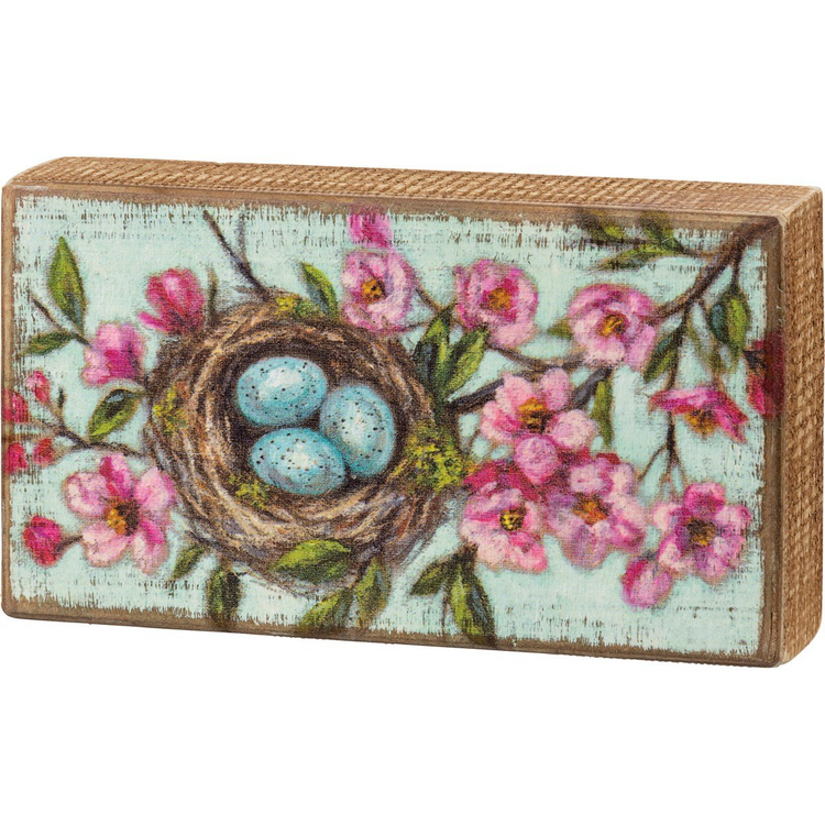Rectangular wooden box sign with a distressed blue background featuring a bird's nest with three blue eggs and pink blossoms.