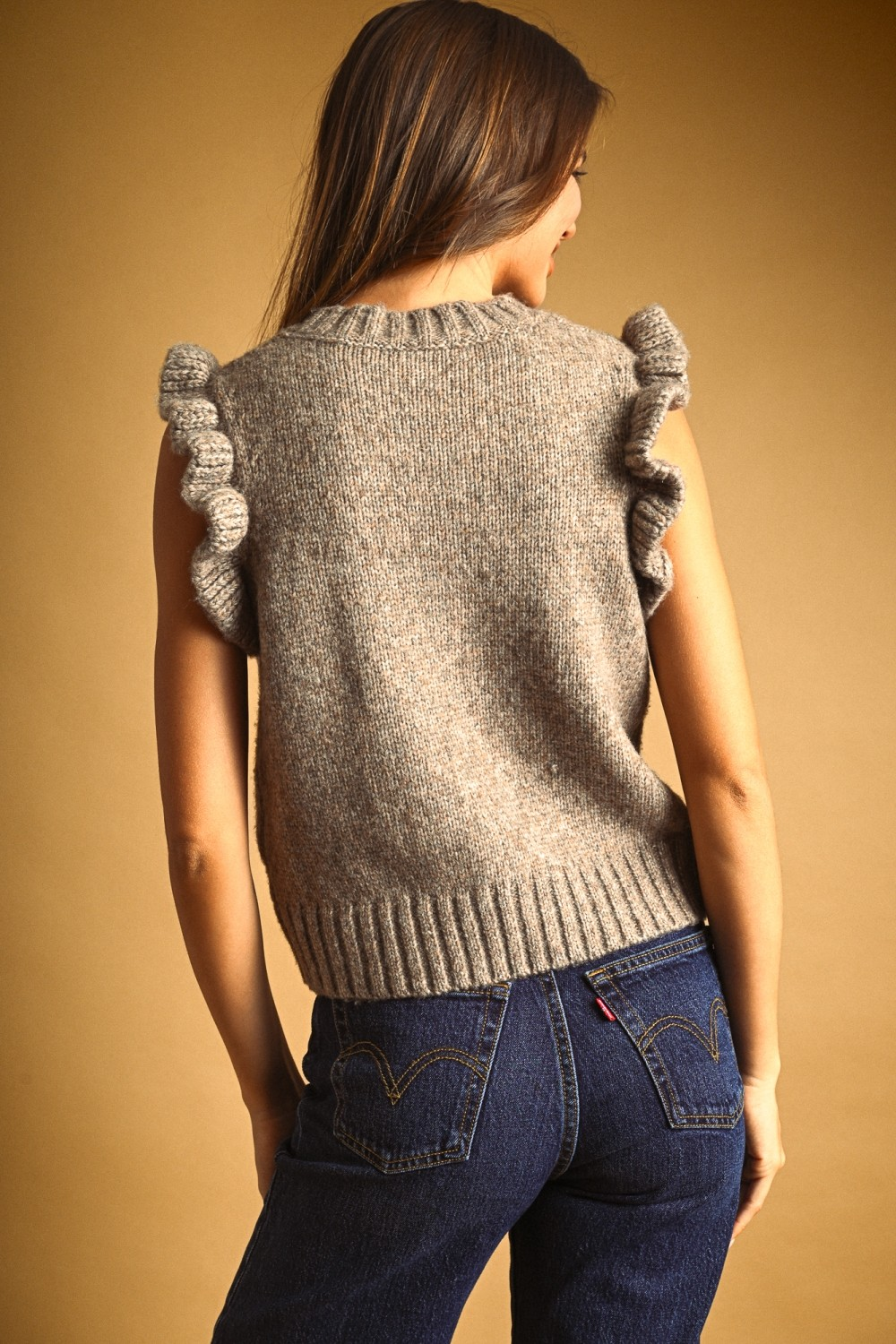 Back view of a woman wearing a heathered grey sleeveless sweater with ruffled armholes and jeans.