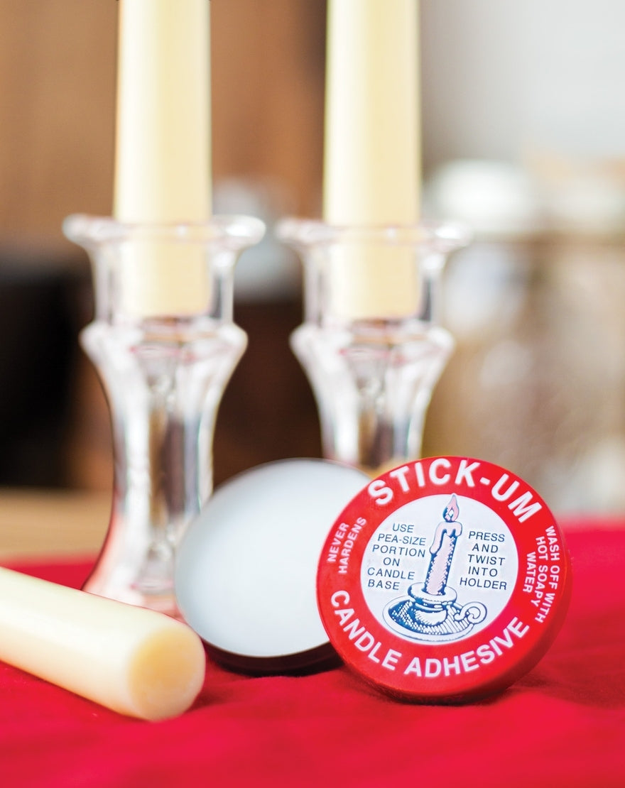 Fox Run Stick-Um Candle Adhesive tin next to white candles in glass holders on a red surface.