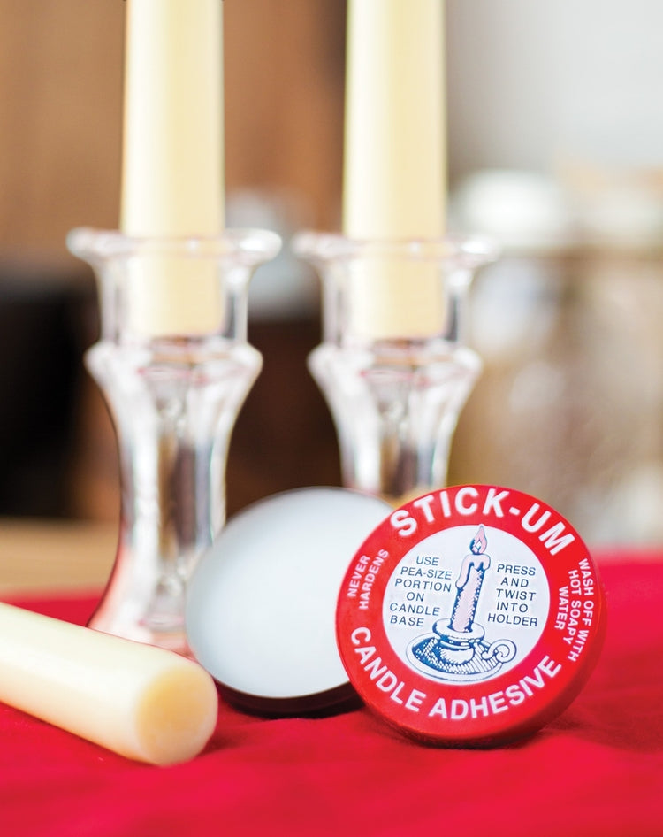 Fox Run Stick-Um Candle Adhesive tin next to white candles in glass holders on a red surface.