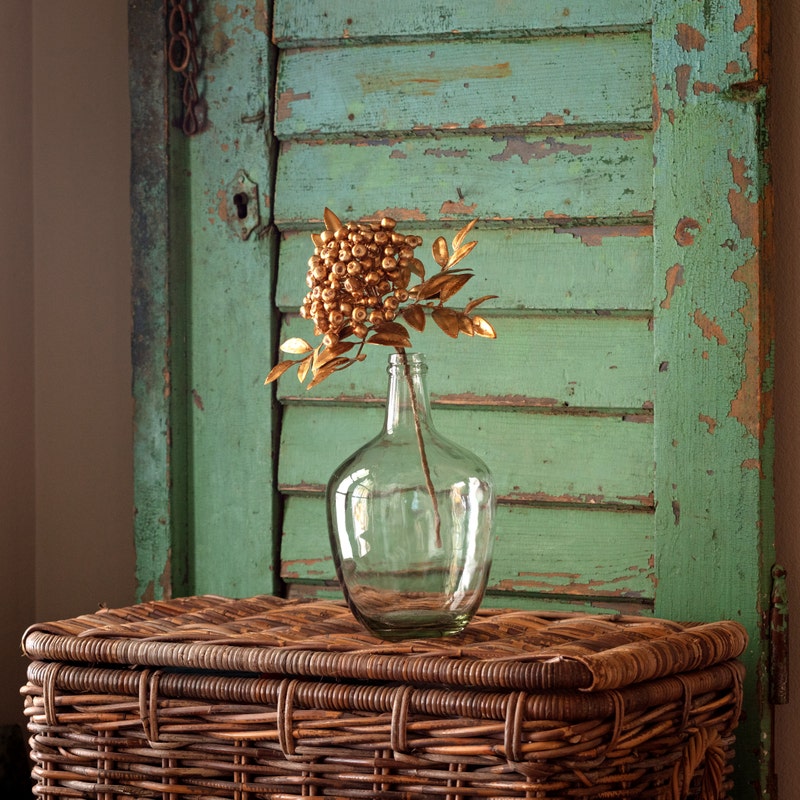 Gold Berry Bunch Metallic faux floral stem in a clear glass vase on a wicker chest.