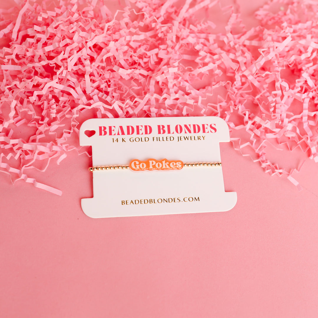 Go Pokes orange and white script charm on a gold beaded bracelet, displayed on a white card.