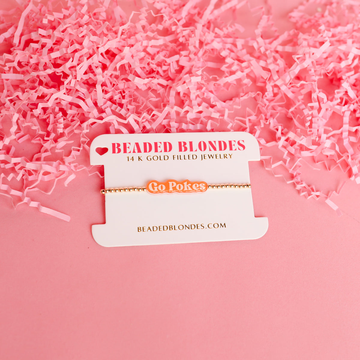 Go Pokes orange and white script charm on a gold beaded bracelet, displayed on a white card.