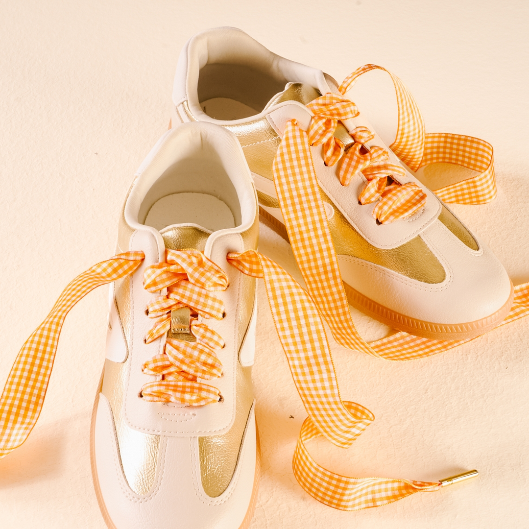 Pair of gold and cream sneakers featuring orange and white gingham ribbon shoelaces.