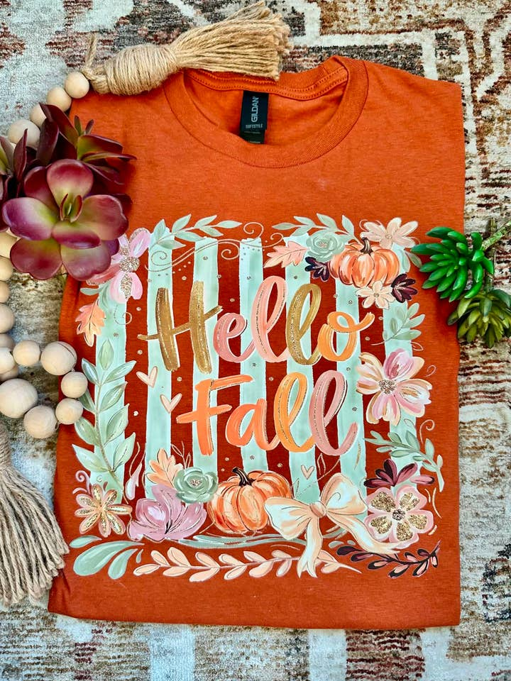 Rust-colored graphic tee with 'Hello Fall' text, pumpkins, and floral accents on a striped background
