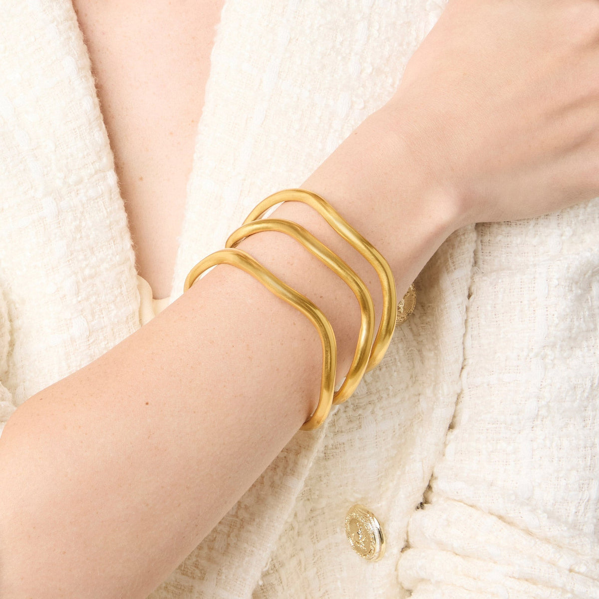 A person wearing three stacked 24K gold-plated Wave Bangles on their wrist with a white tweed jacket.
