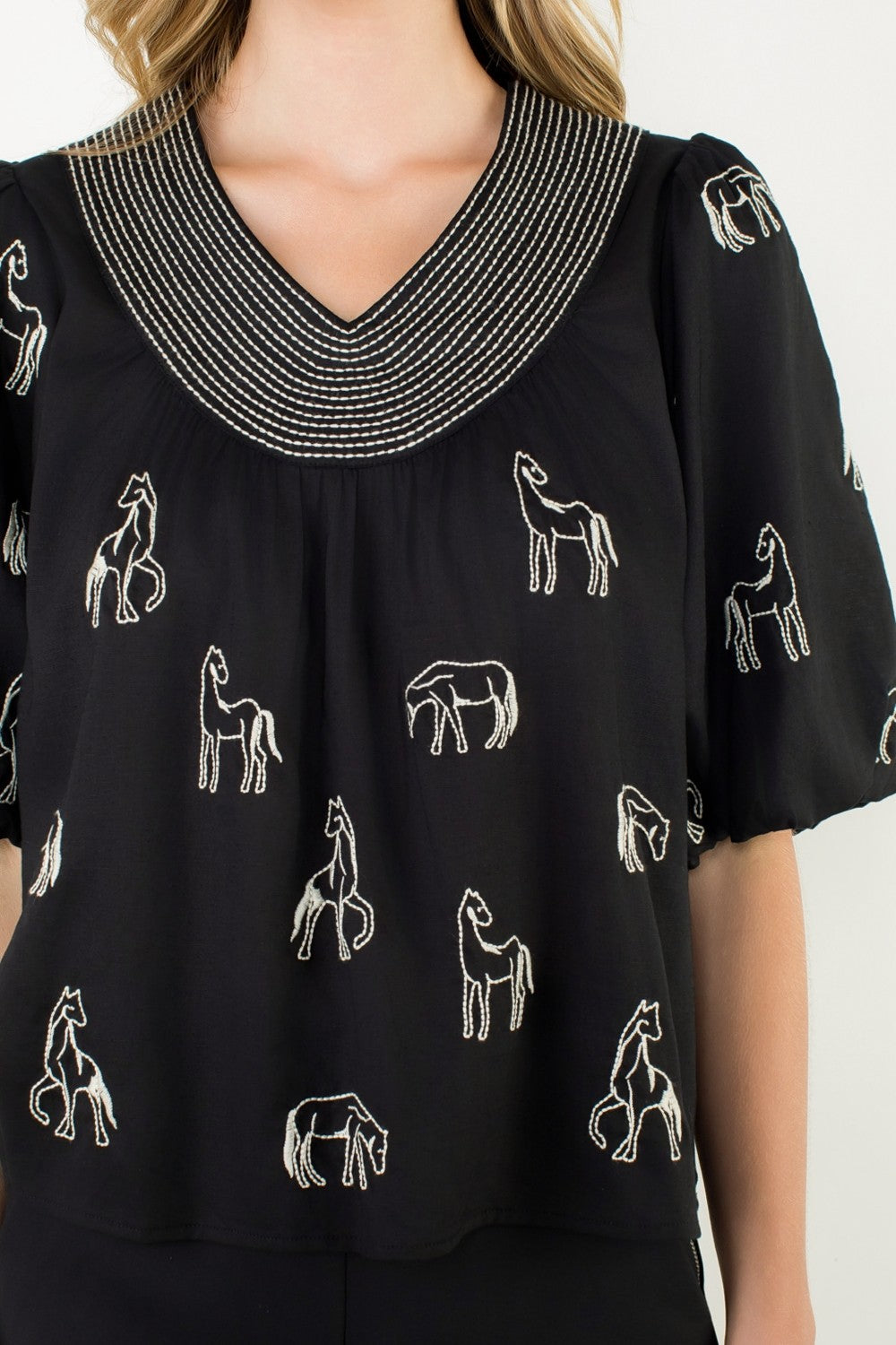 Black V-neck top with white embroidered horses, striped neckline, and puff sleeves.
