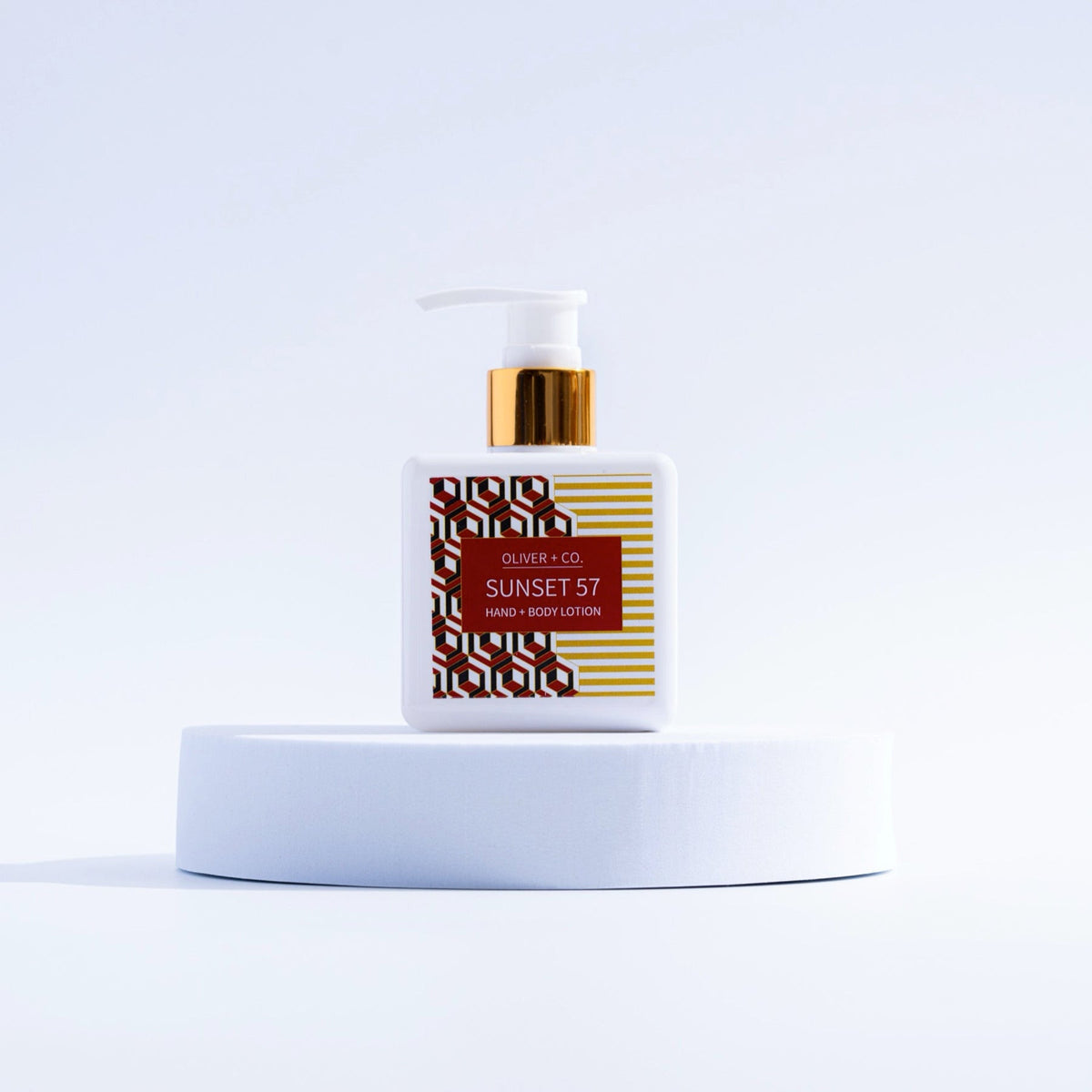 Oliver + Co. Sunset 57 Hand + Body Lotion in a white square bottle with a gold pump on a pedestal.