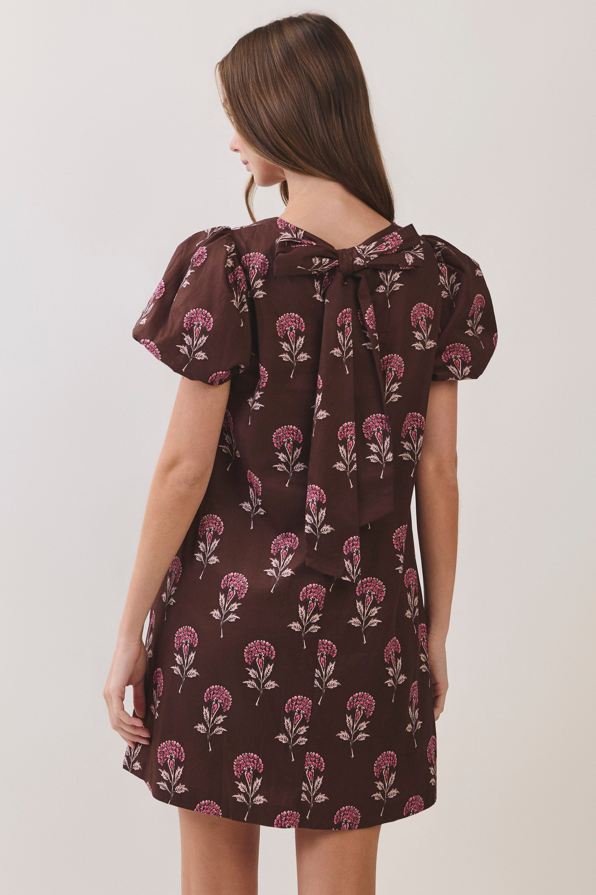 Back view of a brown floral mini dress with short puff sleeves and a large self-tie bow detail.
