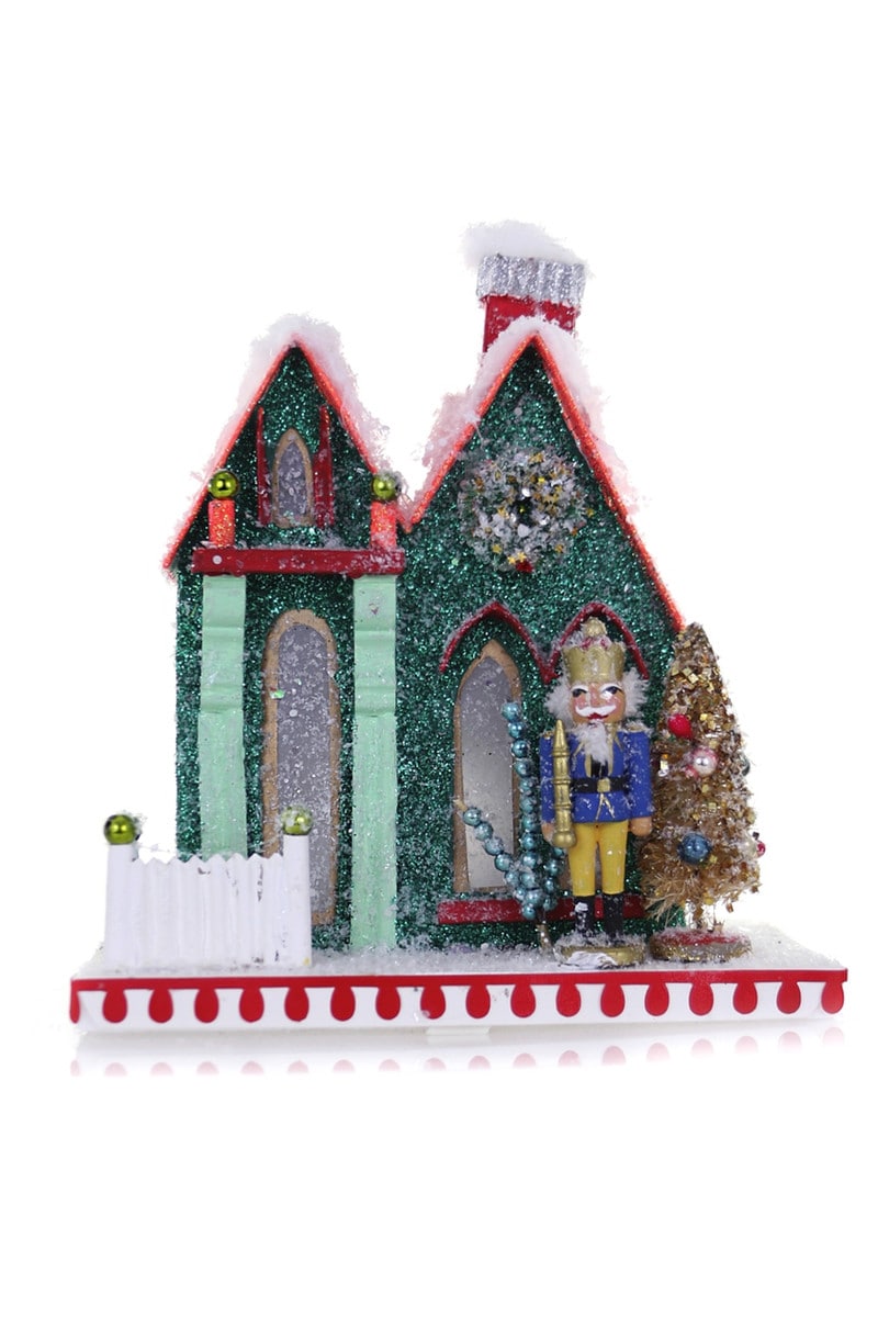 Glittering green cardboard nutcracker cottage with red trim, snow, a nutcracker figure, and a Christmas tree.