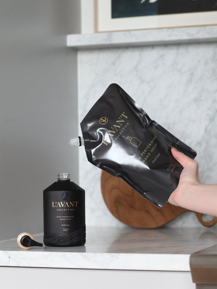 A hand pouring a 32 oz L'AVANT Collective Fresh Linen hand soap refill pouch into a black bottle.