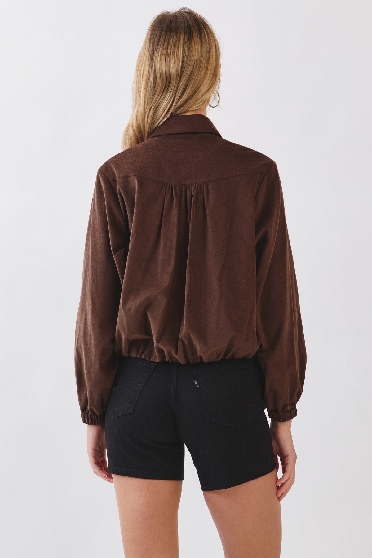 Back view of a woman wearing a brown corduroy zip-up collar jacket with black shorts.